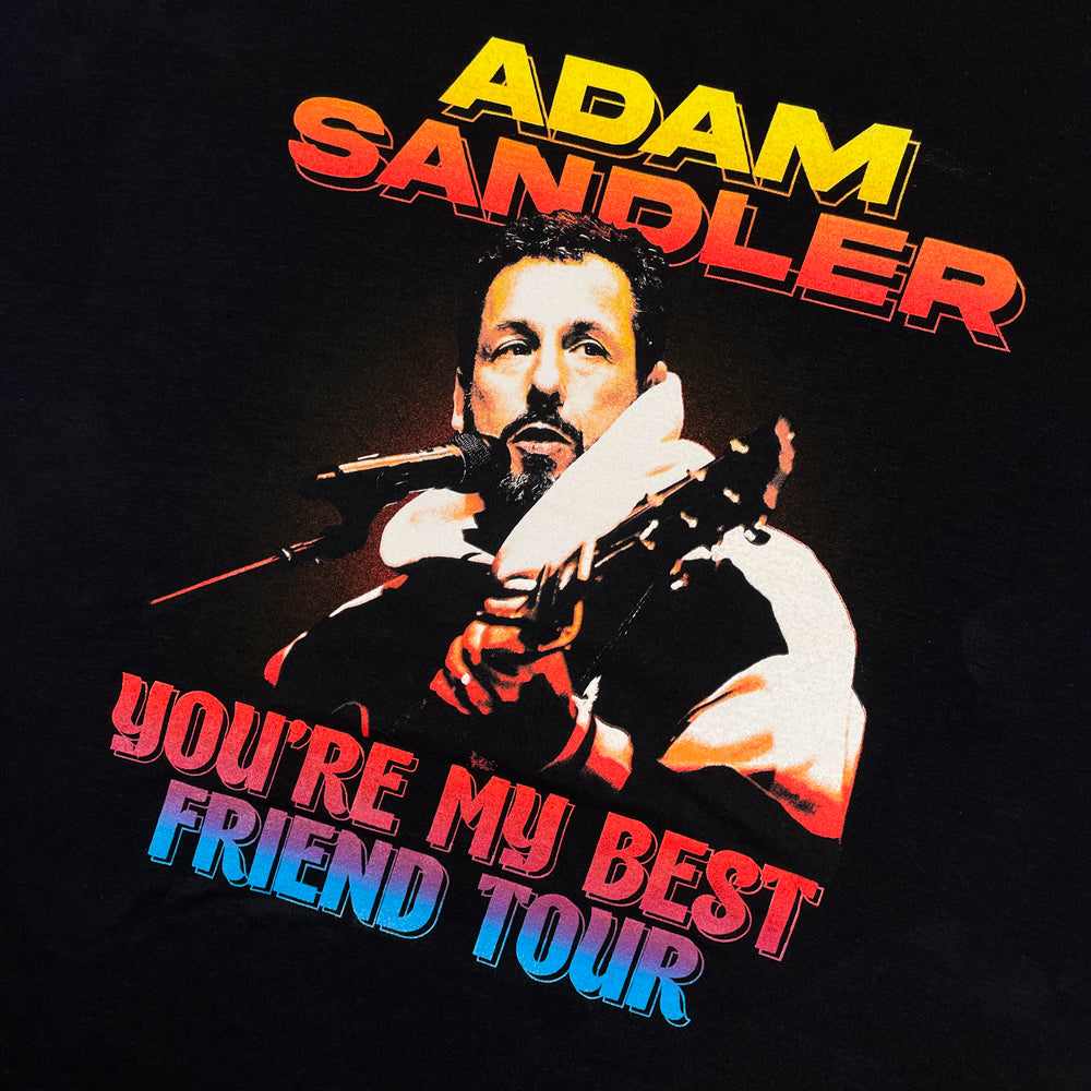 Close up of a black t-shirt depicting Adam Sandler holding a guitar surrounded by text reading "Adam Sandler You're My Best Friend Tour" against white background