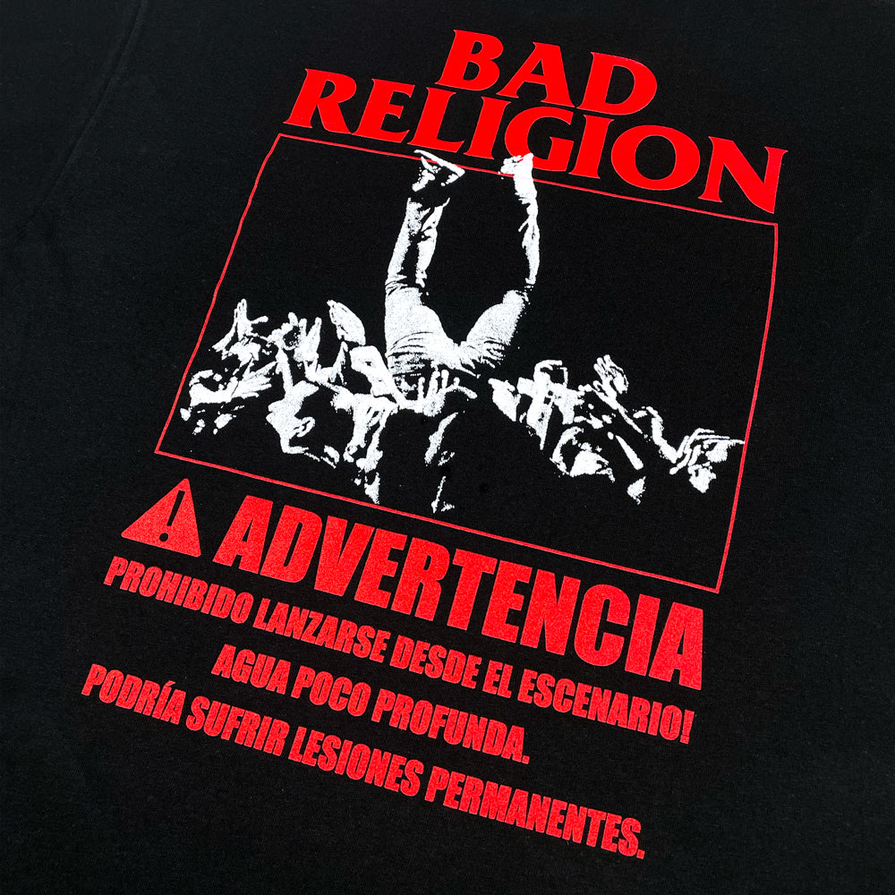 Close up of the back of a black tee with red text reading "Bad Religion, Advertencia, Prohibido Lanzarse Desde El Escenario! Agua Poco Profunda, Podría Sufrir Lesones Permanentes." With a white graphic of a person crowd surfing.