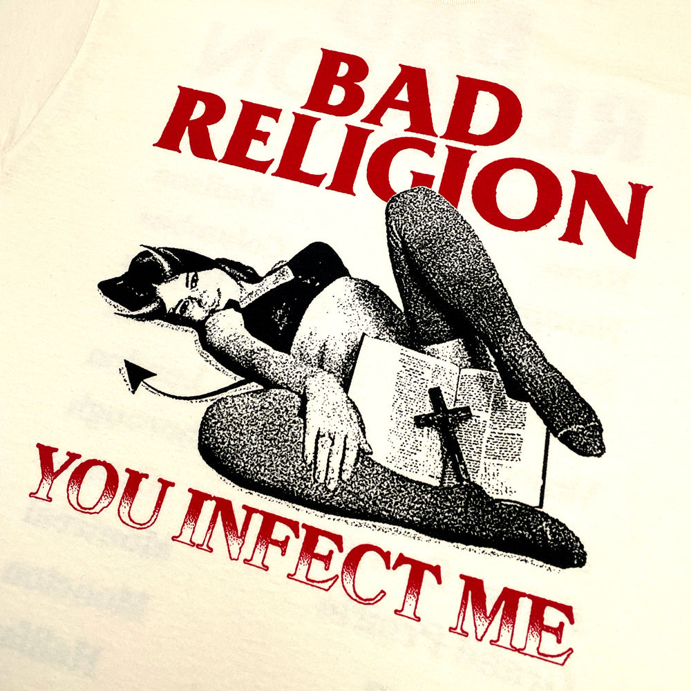 Close up of a cream t-shirt with graphic of a woman with devil horns holding a bible and cross surrounded by text reading "BAD RELIGION YOU INFECT ME"