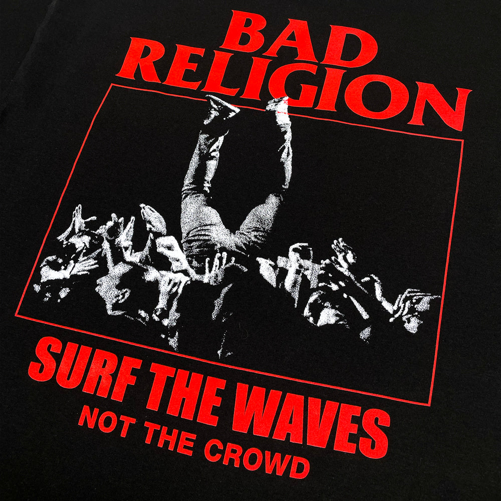 A close up of the back of a black t shirt against a white background with red text reading "Bad Religion, Surf the Waves, Not the Crowd" and a black and white image of a person crowd surfing.