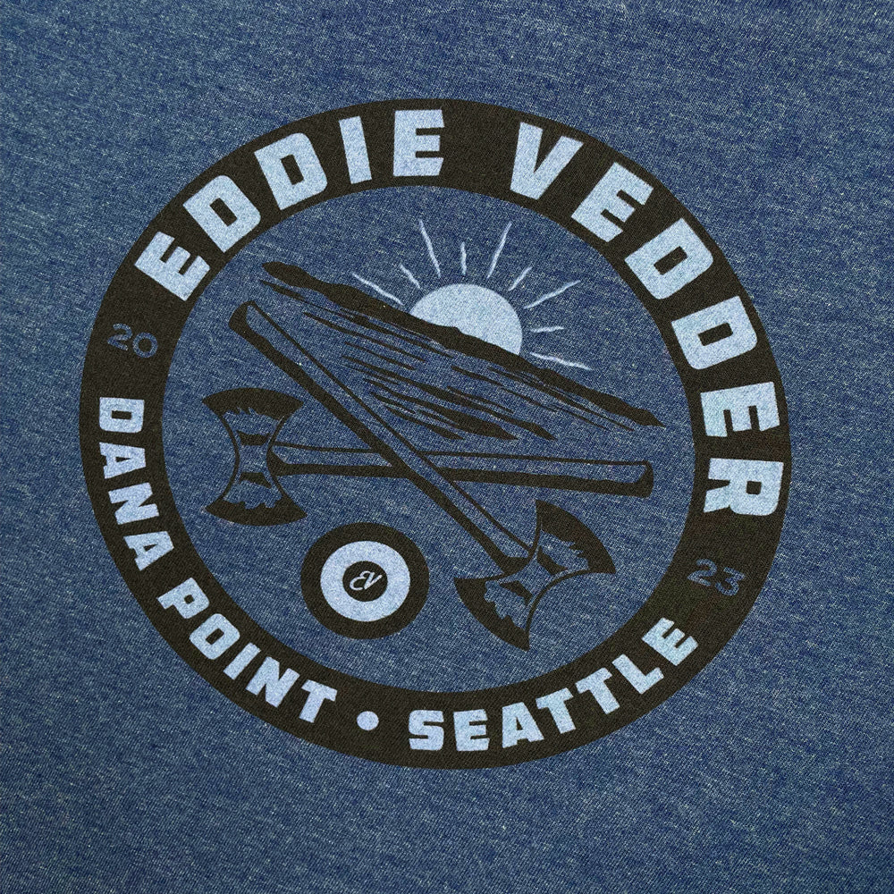 Close up of a heather denim tee with a light blue/black circular design with light blue text reading "Eddie Vedder Dana Point -Seattle 2023" Inside the circle is a light blue sunset with black ocean ripples. There is a design of a pair of axes crossed. Below the axes is a light blue and black target with light blue  letters "EV" in the center.