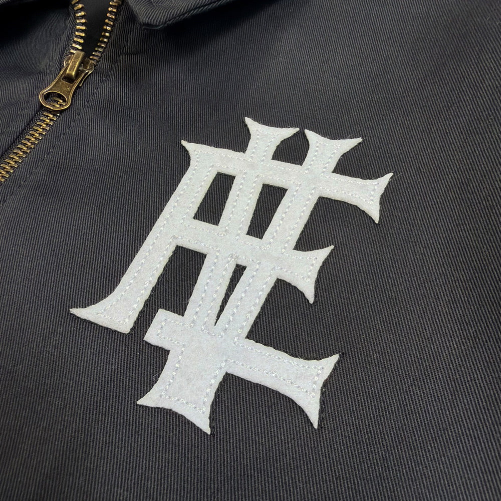 Close-up of white stitched monogram beside the metal zipper on black Eddie Vedder Dana Point Work Jacket