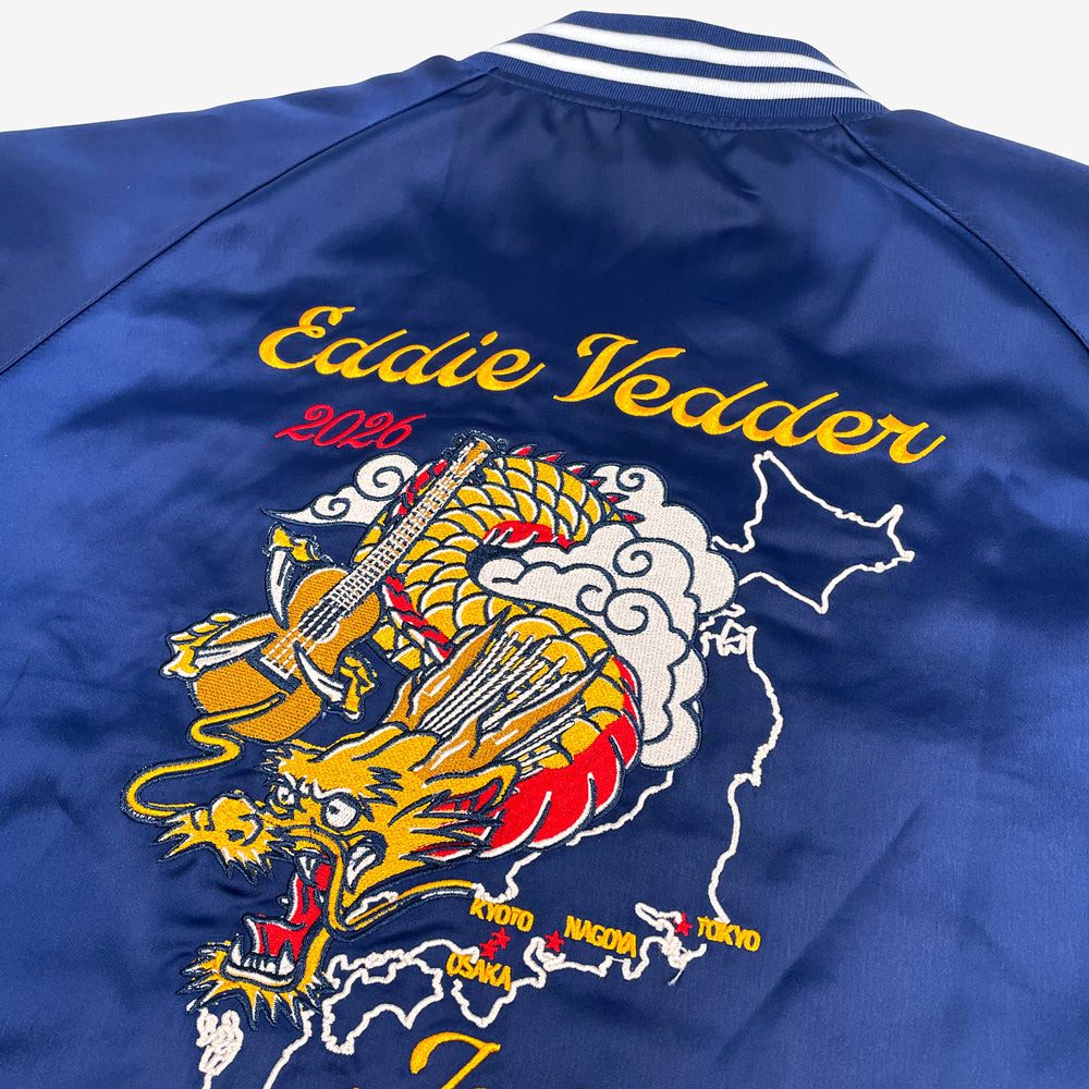 Close up of the back of a navy jacket with white stripes on the collar, cuff, and hip against a white background. Centered on the back is a yellow, orange, and red dragon holding a guitar. the dragon is flying in the clouds and behind the dragon is a white outline of Japan. In yellow the names of the cities of the tour are highlighted. In yellow text on top reads "Eddie Vedder" and below there is yellow text that reads "Japan"