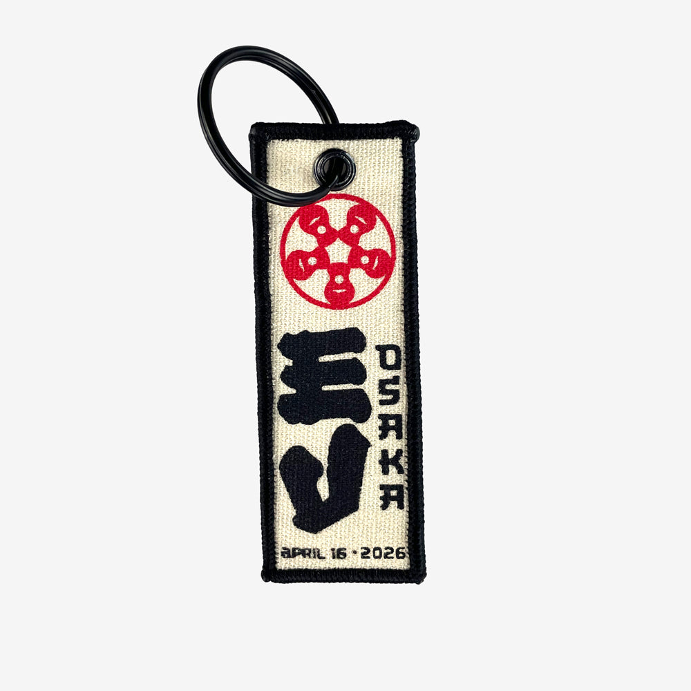 A flight tag keychain against a white background. At the top of the flight tag is a red circular design. In the middle is large black text reading "EV" and to the right of that is black text reading "Osaka" At the bottom in black text there is a date that reads "April 16, 2026"