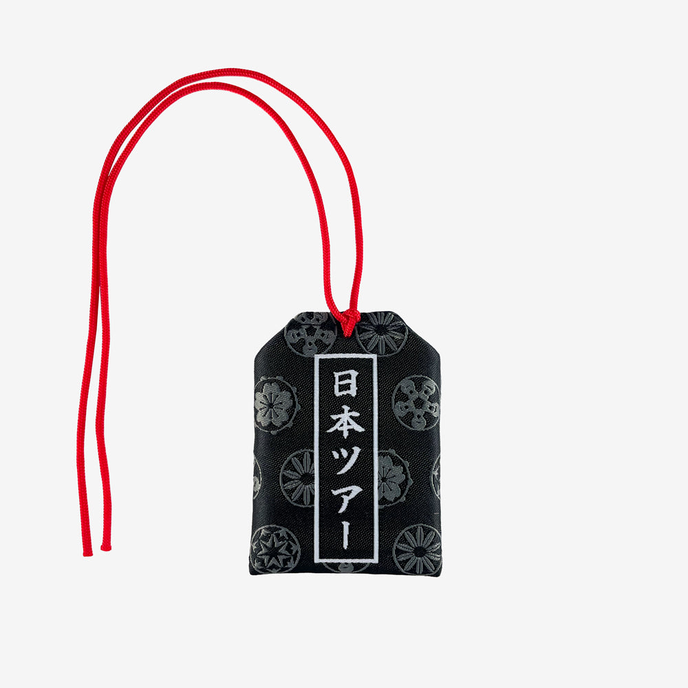 A black amulet bag with red strings against a white background. The Red strings are tied in an ornate knot and there is a white design of a person balancing on top of Mt. Fuji, where there is "Japan" written across the bottom. On the bag are a series of grey circular designs