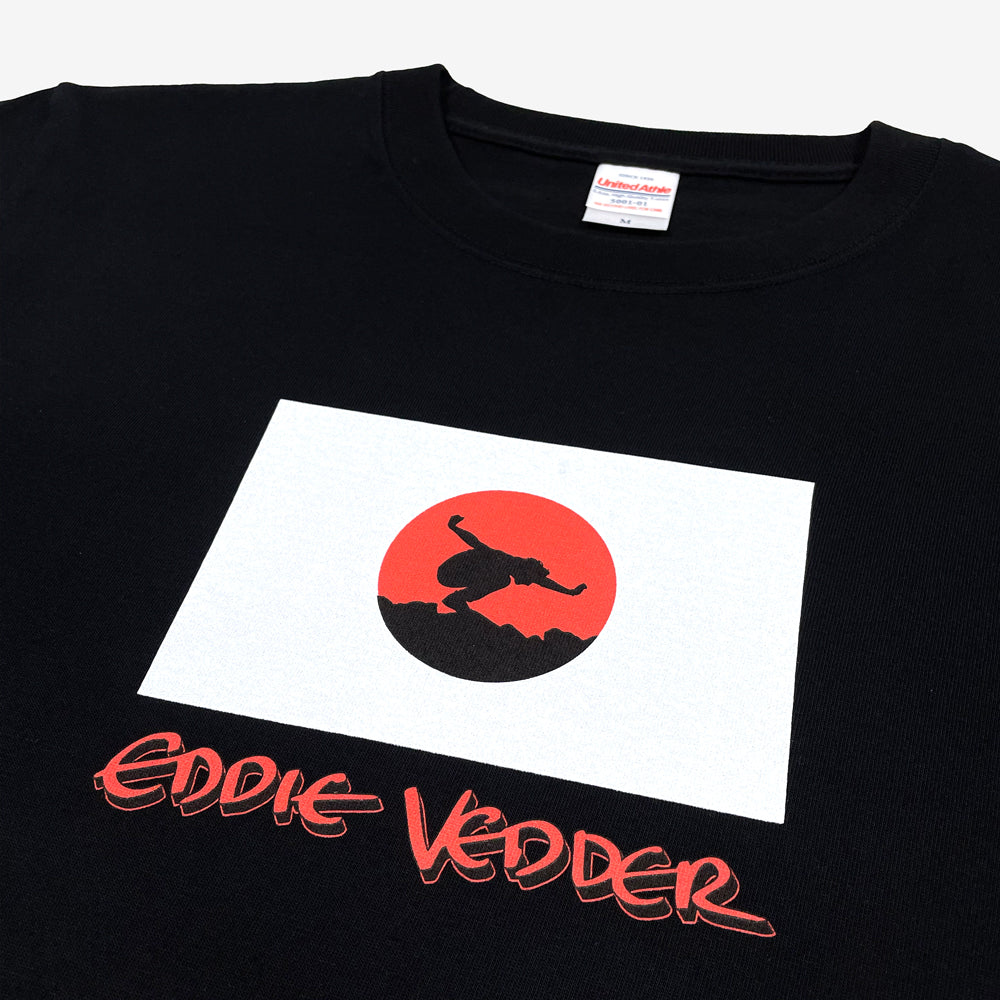 Close up of a black T shirt against a white background with a design of Japanese flag and a silhouette of a person posing in the center. Below the flag design in a red text design that reads "Eddie Vedder"