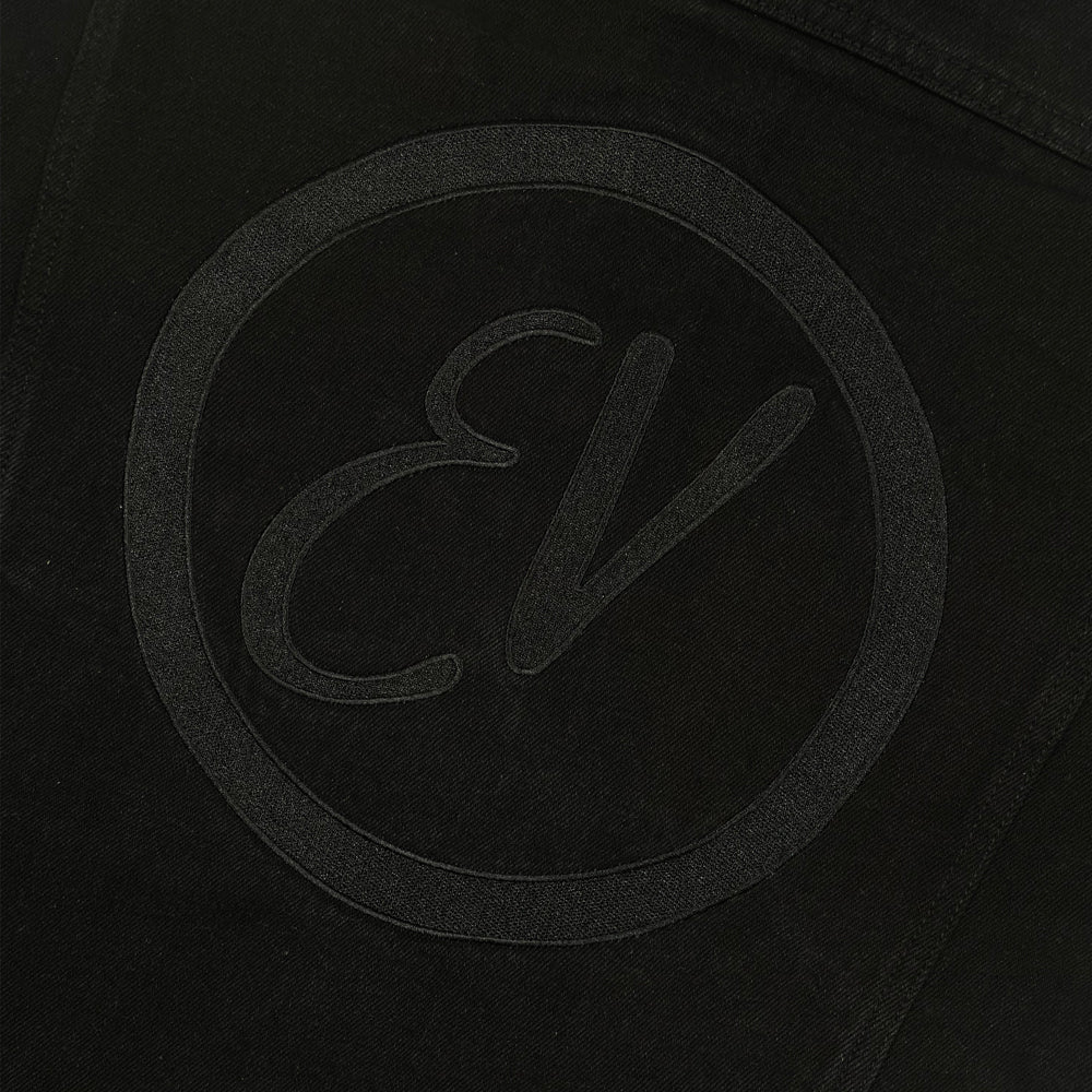 Close up of the back of a black denim jacket with a hoodie against a white background with a black embroidered circular patch the black embroidered text reading "EV"
