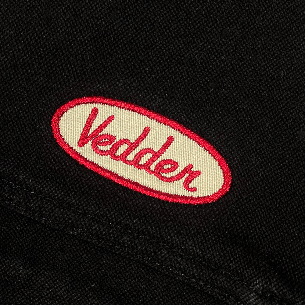 Close up of a black denim jacket with a hood against a white background with a white and red embroidered patch on the chest with text reading "Vedder"