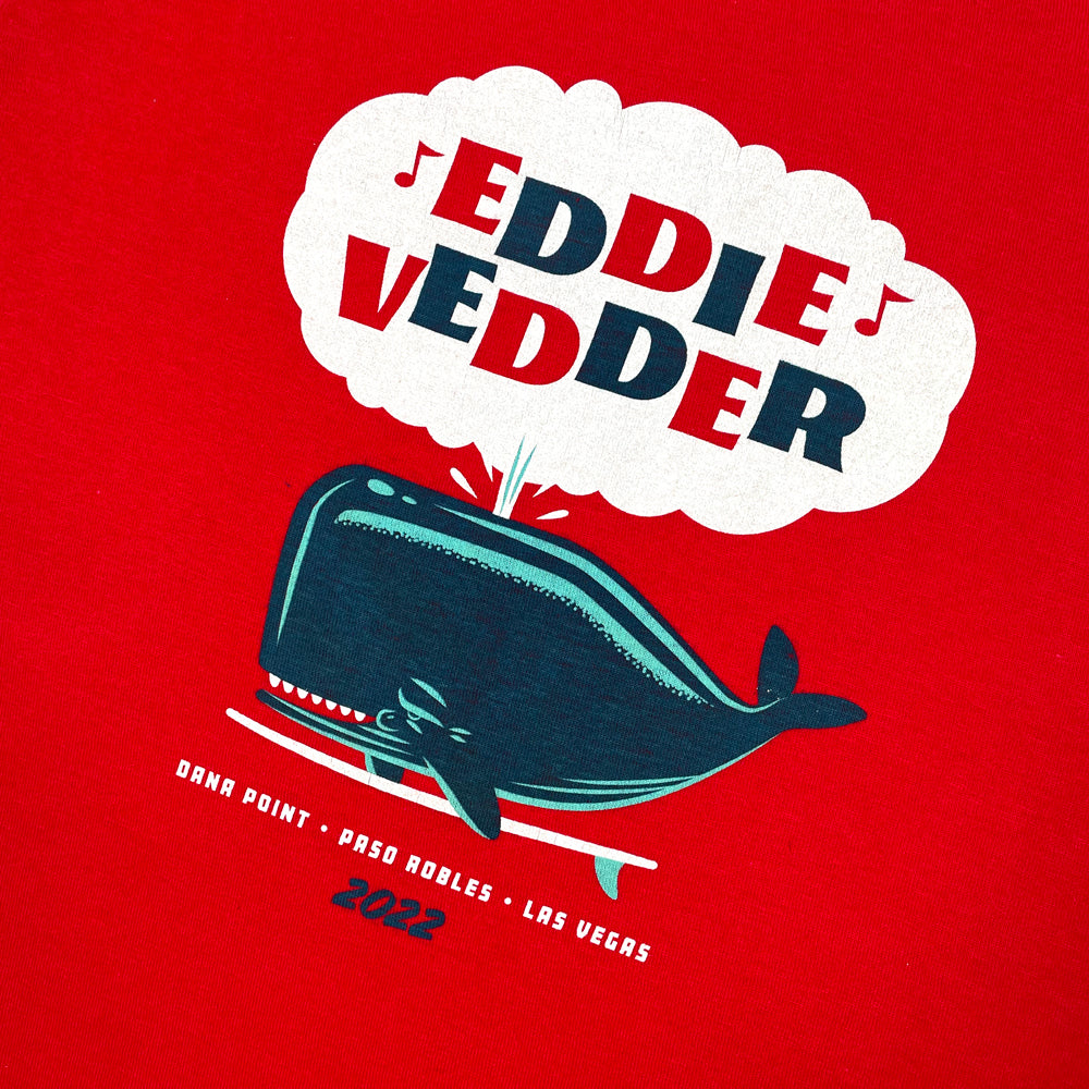 Red kids tee against a white background with a design of a whale on a surfboard shooting out water. Inside the water cloud is alternating red and blue text reading "Eddie Vedder" with music notes on each side of "Eddie." Below the whale is white text reading "Dana Point, Paso Robles, Las Vegas" and below the white text are blue numbers reading "2022"