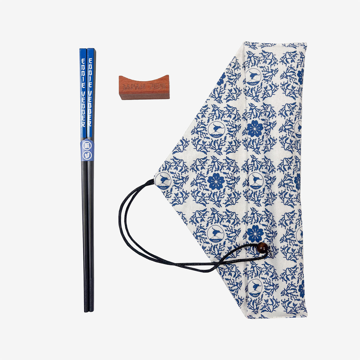 A pair of chopsticks, a wooden rest, and a white cloth bag against a white background. The chopsticks are black with blue ends with white text reading "Eddie Vedder." The cloth bag has a blue floral pattern on it as well as a pattern of a circular design showing a man balancing on top of Mt. Fuji.