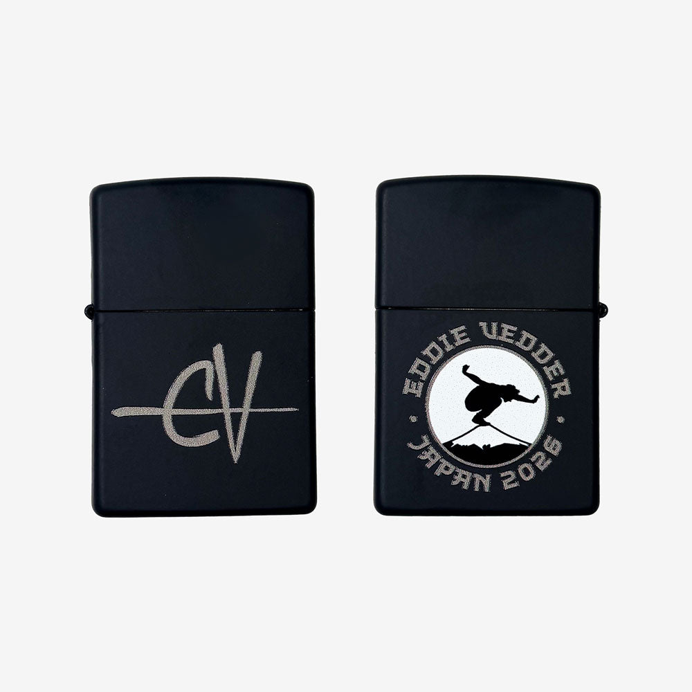 Front and back of a black zippo lighter against a white background. One side has customized "EV" text in silver. The other side has a white circular design of a man balancing on Mt. Fuji. Around the design in silver text reads "Eddie Vedder Japan 2026"