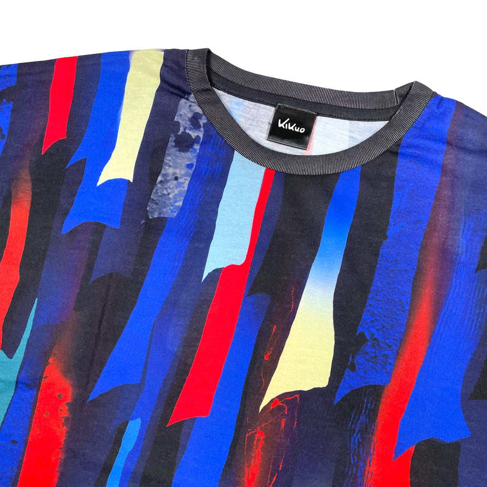 A close-up of a crewneck t-shirt against a white background featuring a bold abstract vertical stripe pattern in blue, red, yellow, and black. The shirt has a dark gray ribbed collar, and a black neck label with the name 'Kikuo' is visible