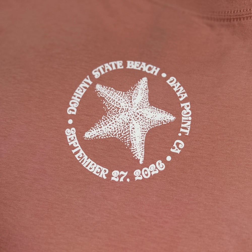 Close-up of white starfish and Doheny State Beach event graphic on desert pink tee