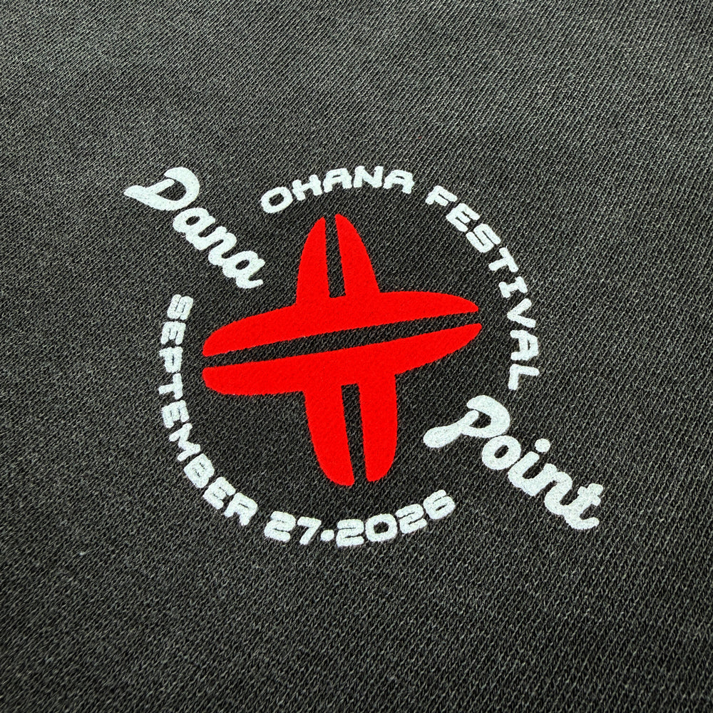 Close up of a  dark gray crewneck sweatshirt with a small circular graphic on the left chest featuring red crossed surfboards and white text that reads "Ohana Festival, Dana Point, September 27, 2026"