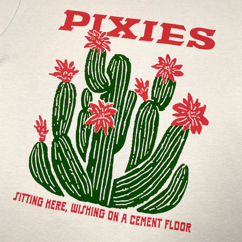 Close up of a beige T-Shirt with green and pink graphic displaying a cactus. Pink lettering reads "Pixies, sitting here, wishing on a cement floor"