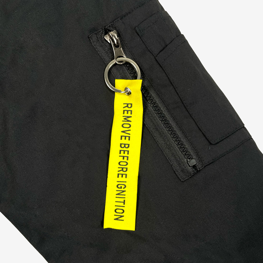 Close up of a black bomber jacket sleeve against a white background with a yellow tag on the sleeve with black text reading "Remove Before Ignition."