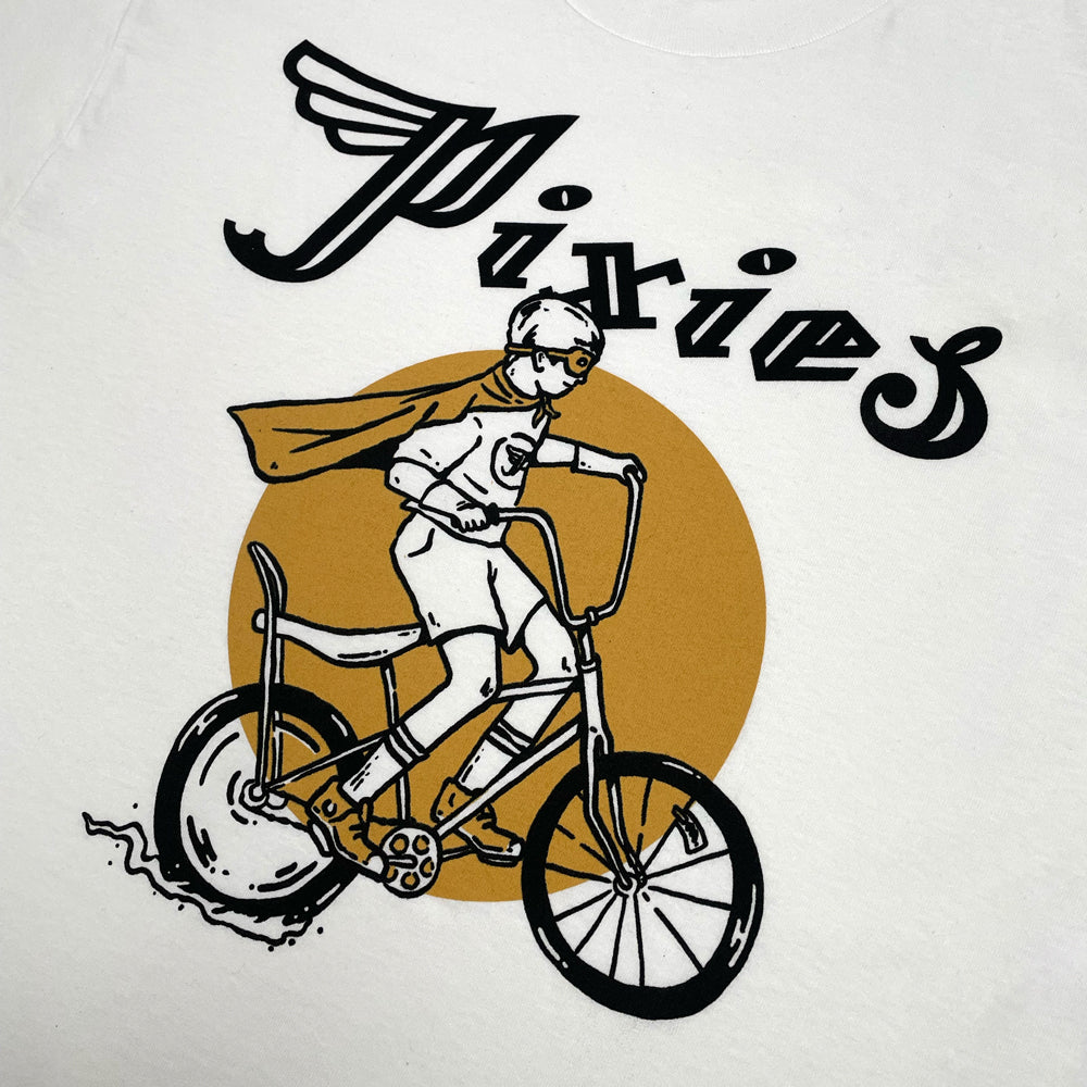 Close up of a white t shirt with a black text logo reading "Pixies." Below the logo is a design of a person dressed in a cape and helmet doing a wheelie on a bike. behind the design is a golden yellow circle.
