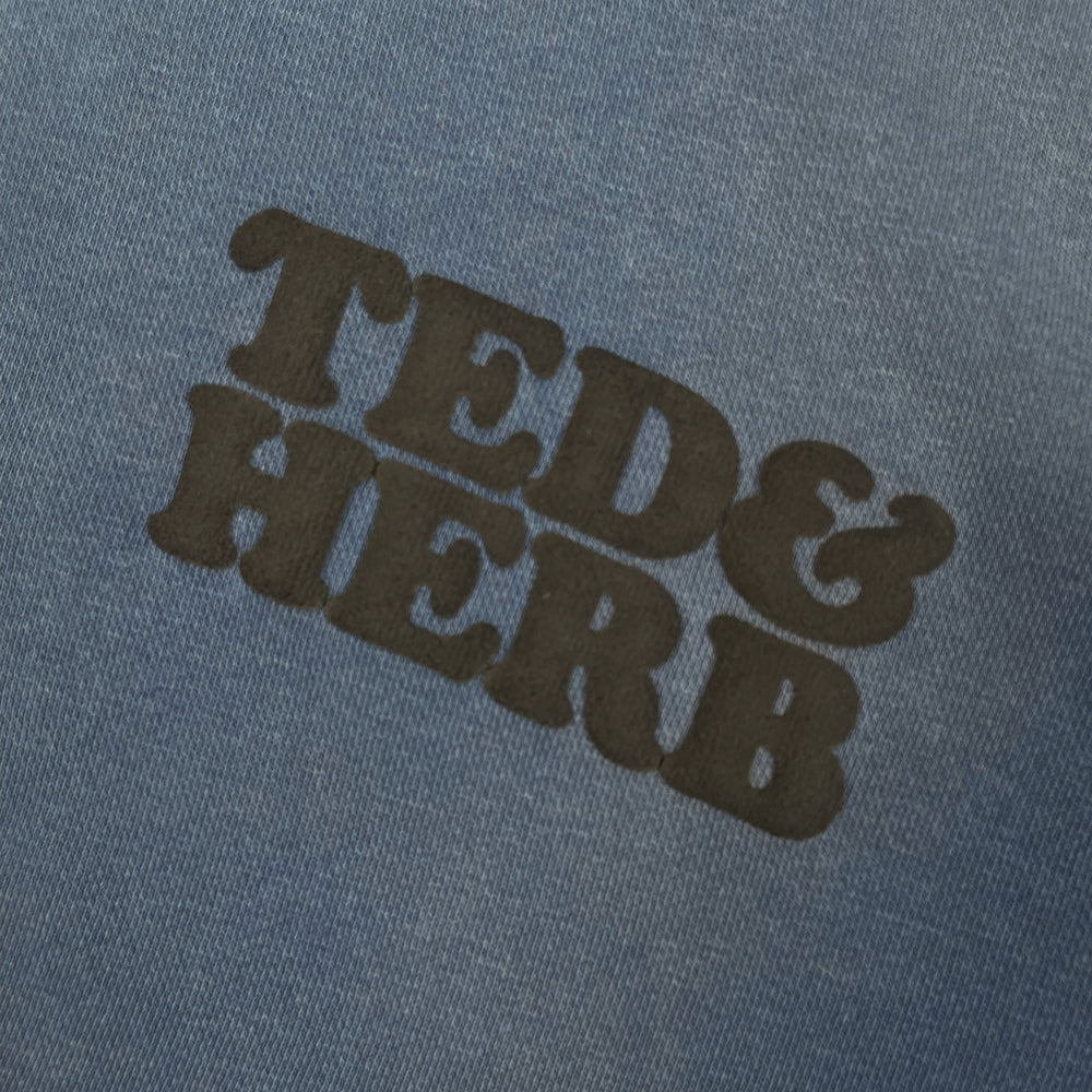 Close up of a blue pullover with black lettering reading "Ted & Herb" against a white background
