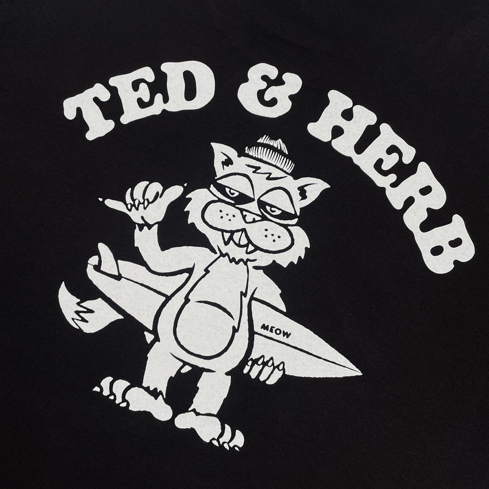 Close up of a black T shirt with white arching text reading "Ted & Herb" and a white design of a cat holding a surf board below.